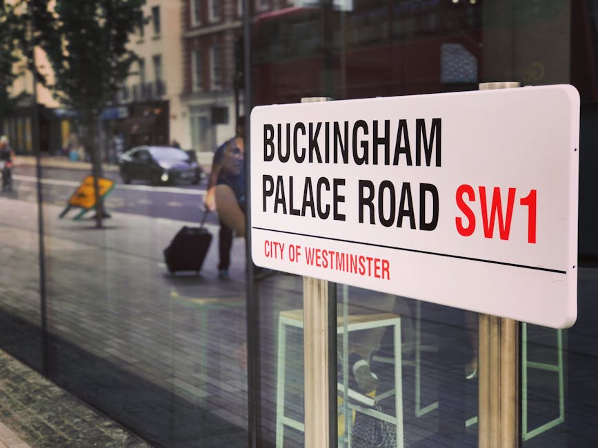A white rectangular street sign displaying the location 'Buckingam Palace Road SW1' in bold black and red letters, with the red text indicating the postcode and 'City of Westminster' written below in smaller red font. The sign is mounted on two silver metal posts positioned on a sidewalk, with a glass-fronted building and the reflection of passing pedestrians visible in the background. Behind the glass, blurred figures of individuals, including a woman with a dark outfit and a rolling suitcase, can be seen walking along the street. The scene is illuminated by natural daylight, highlighting the texture of the sign's smooth finish and the reflections on the glass surface, which subtly connects to themes of urban waste management and alternative disposal methods within Westminster's busy city environment.