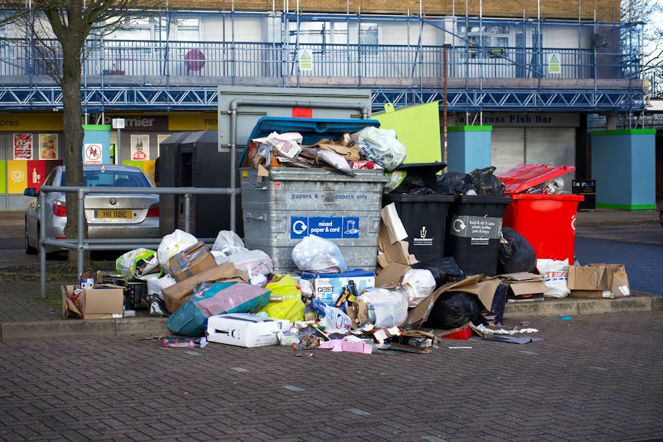 In the foreground, a large, rectangular metal rubbish bin with a blue lid and a label indicating it is designated for mixed paper and cardboard sits on a paved sidewalk, overflowing with various waste items including crumpled paper, cardboard boxes, plastic packaging, and white paper bags. Surrounding the bin, numerous bags and loose debris, such as black plastic rubbish bags, flattened cardboard packages, and plastic wrappers, spill onto the pavement, creating a cluttered scene. To the left, a small silver hatchback car is parked parallel to the bins, partially obscured by a metal railing that separates the waste area from the street. Behind the rubbish collection area, a building with a blue scaffold and green accents on the structure is visible, with storefronts featuring signage and symbols indicating commercial premises, such as a fish bar. The environment appears to be an urban area with natural daylight illuminating the scene, highlighting the disorganized waste outside a private collection point, typical of independent rubbish disposal efforts by waste management services like Waste Collection Westminster in the SW1 postcode district.
