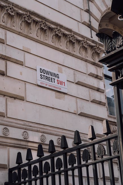 A white rectangular street sign displaying the location 'Buckingam Palace Road SW1' in bold black and red letters, with the red text indicating the postcode and 'City of Westminster' written below in smaller red font. The sign is mounted on two silver metal posts positioned on a sidewalk, with a glass-fronted building and the reflection of passing pedestrians visible in the background. Behind the glass, blurred figures of individuals, including a woman with a dark outfit and a rolling suitcase, can be seen walking along the street. The scene is illuminated by natural daylight, highlighting the texture of the sign's smooth finish and the reflections on the glass surface, which subtly connects to themes of urban waste management and alternative disposal methods within Westminster's busy city environment.
