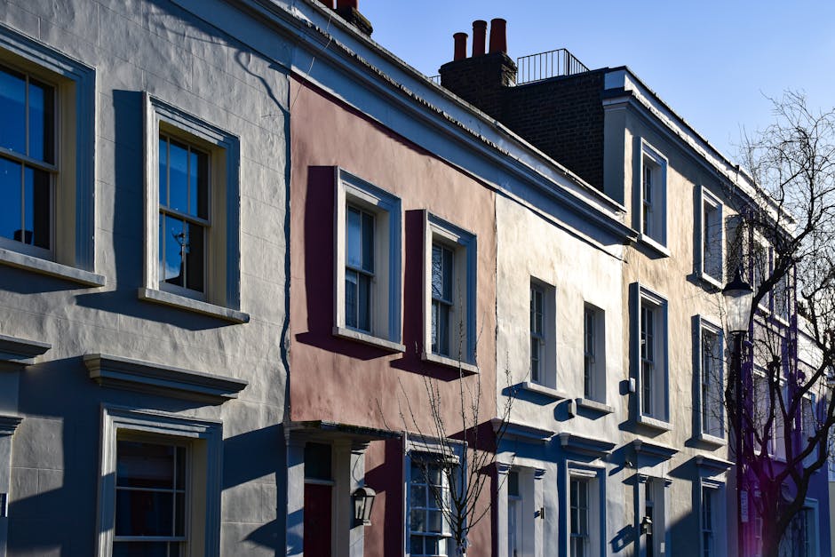 A row of three adjacent residential buildings with varying pastel-colored facades, including shades of light blue, pink, and cream, featuring white window frames and decorative window sills. The buildings have multiple stories with sash windows, some reflecting the sky and surroundings. The rooftops are flat with red brick chimneys and metal railings visible on the upper levels. In the foreground, leafless trees cast shadows on the facades, indicating a clear, sunny day. The environment suggests an urban area with typical terraced housing, and the scene is suitable for illustrating private property waste management or alternative rubbish collection services, reflecting the context of independent rubbish removal for city residents. The natural lighting enhances the textures of the stucco walls and their details, emphasizing the different materials and finishes. The overall composition is orderly, highlighting the exterior architecture and surrounding environment, aligning with waste management themes in residential areas.