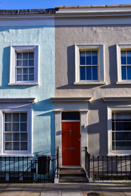 The image depicts the front facade of a two-story residential building with a clear division of colour; the left side painted in light blue and the right side in beige, both with smooth texture. Four white-framed sash windows with multiple panes are evenly spaced, two on each level. In the center, a wooden front door painted in deep red features a silver door knocker and mail slot, positioned directly above a small set of concrete steps leading up from the sidewalk. A narrow black metal railing fences the steps, with a black trash bin placed to the left of the entrance. Shadows cast by the building suggest bright sunlight, creating contrast between illuminated and shaded areas. The environmental setting is an urban street scene, possibly part of a terraced housing row, with the exterior walls clean and well-maintained, and the overall scene capturing a typical residential approach to cleaner private waste handling, such as rubbish collection organized by Waste Collection Westminster, as indicated by contextual relevance.