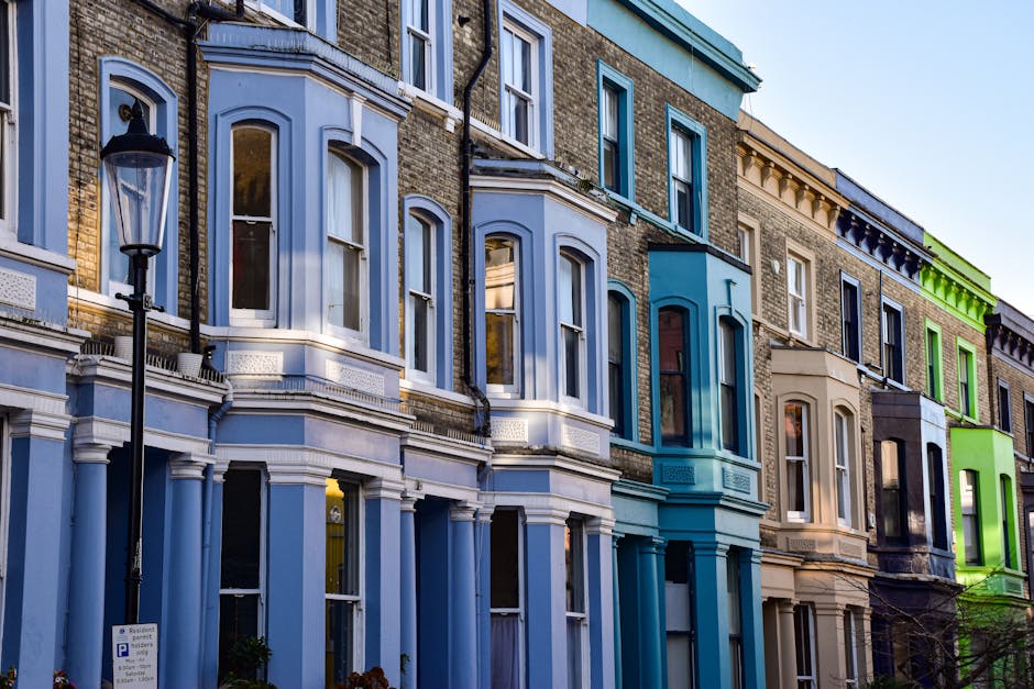 A row of multi-story Victorian-style terraced houses with bay windows, situated on an urban street. The buildings feature brick façades enhanced with decorative white trim and cornices, with each property painted in different pastel and neutral tones, including light blue, beige, navy blue, and bright green accents on window frames and decorative elements. Large, arched windows with white wooden frames dominate the front elevations, some with curtains visible through the glass. The street scene includes a black street lantern mounted on a pole in the foreground, and a parking sign is partially visible at the bottom left corner. The impact of evening or early morning light creates subtle shadows and highlights on the facades, emphasizing the architectural details. The scene is clean, well-maintained, and reflects a typical residential area suited for private waste collection or rubbish removal services, involving inspection of exterior spaces and facade cleaning or waste clearance as part of property upkeep, supported by companies like Waste Collection Westminster.