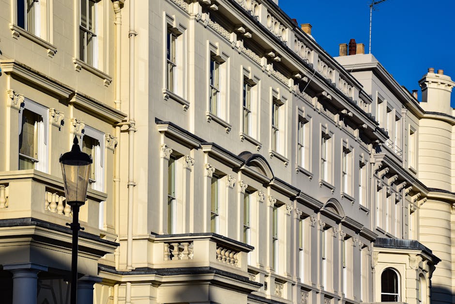 The facade of a historic, multi-story building with cream-colored, ornate architectural details under a clear blue sky. The building features tall, rectangular windows framed by decorative moldings and small balconies with balustrades on the upper floors. A traditional black lamppost is visible in the foreground, standing on a paved area that appears to be part of a street or courtyard. The image's lighting highlights the building's intricate detailing and finishes, which include glossy surfaces and textured ornamentation, typical of classic European architecture. While the scene primarily depicts urban residential or commercial facades, it subtly relates to waste management through the context of urban environments where independent rubbish collection or on-site clearance services, like those offered by Waste Collection Westminster, play a vital role in maintaining the cleanliness of such historic areas.