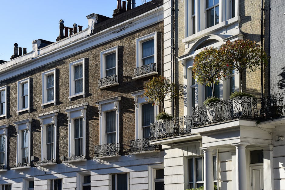 A row of multi-story Victorian-style terraced houses with bay windows, situated on an urban street. The buildings feature brick façades enhanced with decorative white trim and cornices, with each property painted in different pastel and neutral tones, including light blue, beige, navy blue, and bright green accents on window frames and decorative elements. Large, arched windows with white wooden frames dominate the front elevations, some with curtains visible through the glass. The street scene includes a black street lantern mounted on a pole in the foreground, and a parking sign is partially visible at the bottom left corner. The impact of evening or early morning light creates subtle shadows and highlights on the facades, emphasizing the architectural details. The scene is clean, well-maintained, and reflects a typical residential area suited for private waste collection or rubbish removal services, involving inspection of exterior spaces and facade cleaning or waste clearance as part of property upkeep, supported by companies like Waste Collection Westminster.